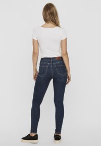 Jean skinny bleu foncé à taille haute, avec une patch en cuir au dos et deux poches arrière. Assorti avec un t-shirt blanc.