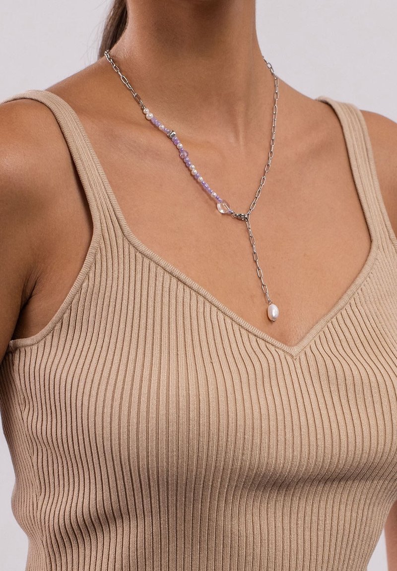 Gros plan d'une femme portant un débardeur côtelé beige et un collier chaîne argenté avec des perles violettes et un pendentif en perle suspendu.