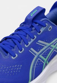 Sininen urheilukenkä, jossa on teksturoitu yläosa, verkkokohdat, heijastavat yksityiskohdat ja vihreät yksityiskohdat. Varustettu perinteisellä nauhoituksella ja kantapääntuen nauhalla.