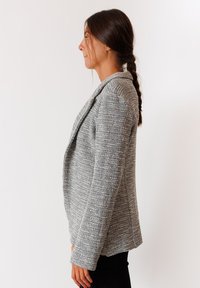 Blazer grigio con una trama testurizzata, frontale aperto e colletto sciallato. Presenta maniche lunghe e una vestibilità su misura, mostrato di lato.