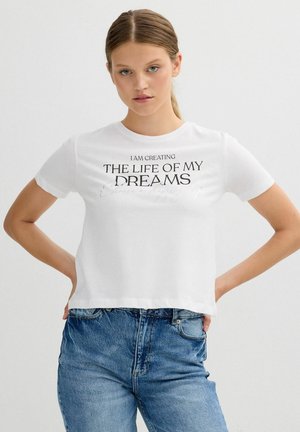 Camiseta de algodón blanca de manga corta, con texto en negro: "ESTOY CREAMDO LA VIDA DE MIS SUEÑOS." Combinada con jeans de talle alto azul.