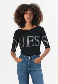 T-shirt nera con lettering in strass che spellano "GUESS" sul frontale; presenta maniche a tre quarti e un ampio scollo.