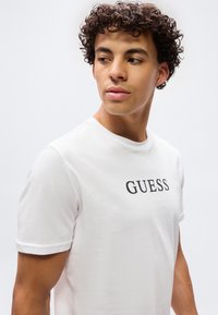 Νεαρός με σγουρά μαλλιά που φοράει λευκό μπλουζάκι με την επιγραφή "GUESS" στο στήθος, κοιτάζοντας προς τα πλάγια μπροστά σε απλό φόντο.