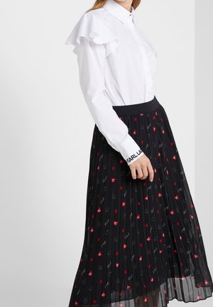 Žena nosící bílou blůzu s volánkovým detailem na ramenou a černou plisovanou sukni s motivem červených symbolů karet a bílého písma.