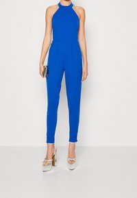 Jumpsuit bleu à col halter en tissu extensible, coupe ajustée avec des plis, associé à des sandales à talons hauts argentées et à une petite pochette ornée.