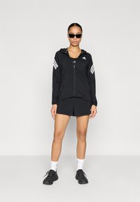 Chaqueta deportiva negra con cremallera, con rayas blancas en las mangas; combinada con pantalones cortos negros y zapatillas negras.