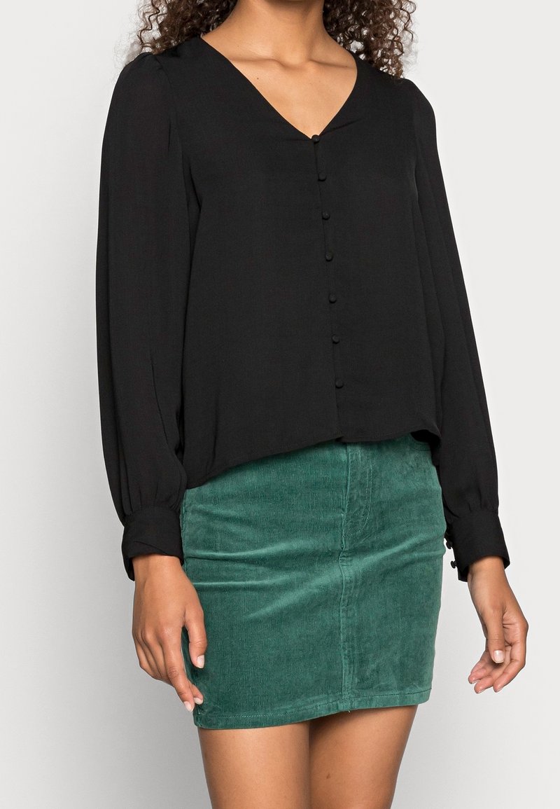 Blouse noire avec un décolleté en V et des boutons sur le devant, associée à une mini-jupe en velours côtelé vert. La blouse possède des manches longues avec des détails de poignets.
