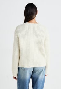 Bélelt, off-white színű, vastag kötésű pulóver, bordás textúrával, leengedett vállakkal és laza szabással. Világoskék, bő szárú farmernadrággal párosítva.