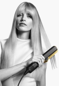 En svart och guld hårsträcka som hålls av en person med långt, rakt blont hår. Handtaget har en distinkt guldaccent och texturerat grepp.