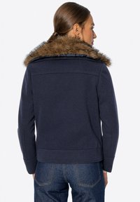 Marineblaue Jacke aus strukturiertem Material, mit einem Kunstfellkragen und tailliertem Schnitt, kombiniert mit dunklen Jeans.