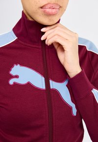 Bordeauxkleurige zip-up trainingsjack met een hoge kraag, voorzien van een blauw en roze Puma-logo en lichtblauwe schouderaccenten. De stof is soepel en gestructureerd.