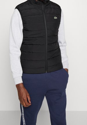 Mann trägt eine schwarze gesteppte Weste mit grünem Krokodil-Logo, hellgraues Langarmshirt und dunkelblaue Hose mit seitlichen Streifen.