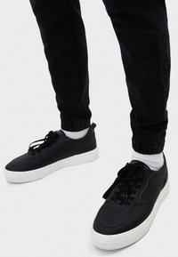 Zapatillas de cuero negras con suelas de goma blancas, punta redonda y cordones planos. Llevadas con pantalones oscuros y calcetas blancas. Diseño sencillo.
