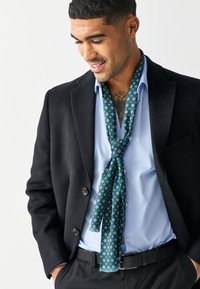Green patterned scarf with small circular designs, made of lightweight fabric, worn loosely over a light blue shirt and black blazer.