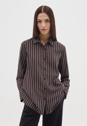 Jonge vrouw met stijlbruin haar draagt een donkerbruine blouse met witte strepen en zwarte broek, staand met handen in de zakken.