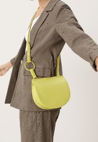 Borsa a tracolla in pelle verde lime con forma curva, tracolla regolabile e dettaglio in metallo circolare, abbinata a una giacca beige perforata.