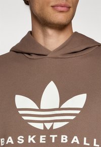 Rudas hoodie su tekstūru, puikuojantis dideliu baltu gėlės grafiku ir žodžiu "KREPŠINIS" ryškiu šriftu.