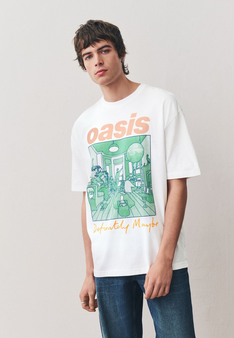 Vit bomulls-t-shirt med en grön grafisk design av ett rum med människor, växter och möbler. "Oasis" och "Definitely Maybe" i orange text.