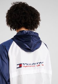 Giacca bianca con dettagli blu navy, dotata di cappuccio e logo "TOMMY SPORT" in rosso e blu sul retro. Materiale elegante e resistente al vento.