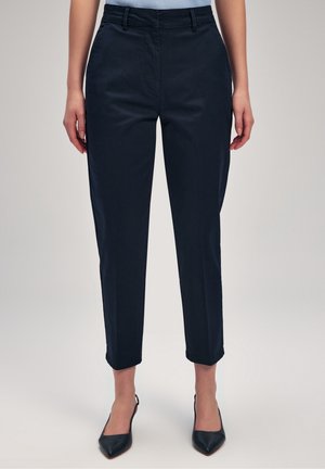 Vrouw met slanke, enkelhoge zwarte broek gecombineerd met zwarte puntige schoenen, staand tegen een effen achtergrond.