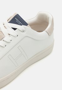 Vita lädersneaker med beige mockadetaljer, strukturerad yta, platta snören och en marinblå logopatch. Har ett subtilt präglat 'H'-mönster.