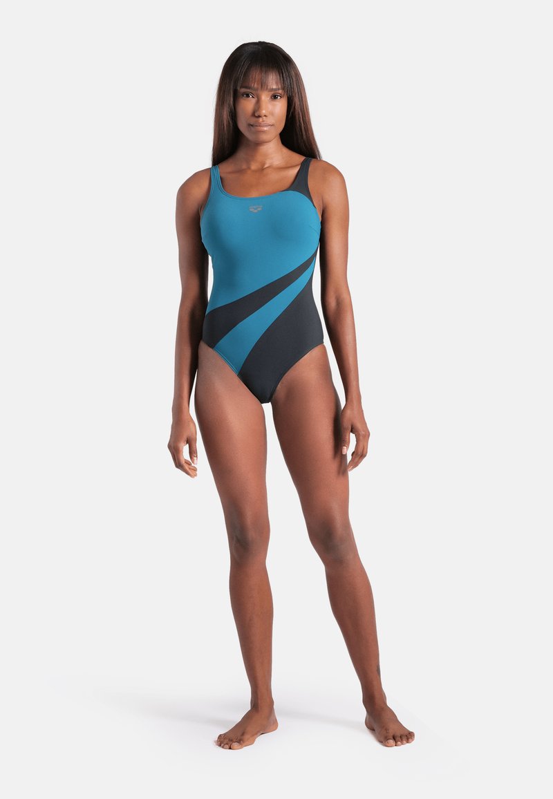 Femme debout pieds nus portant un maillot de bain une pièce sans manches bleu et noir devant un fond blanc uni.