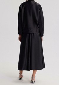 Conjunto de dos piezas en satén negro que incluye una chaqueta entallada con cuello alto y una falda midi fluida. Tacones plateados con tiras completan el conjunto.