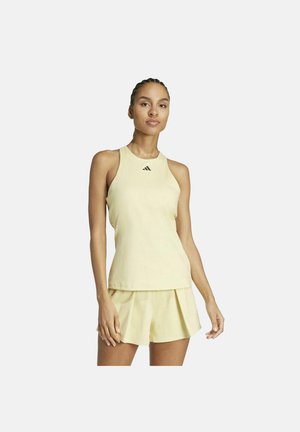 Donna che indossa una canotta sportiva giallo chiaro senza maniche e pantaloncini plissettati abbinati, in piedi davanti a uno sfondo bianco.