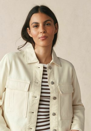 Young woman with dark hair wears a cream buttoned jacket over a brown and white striped shirt, posing against a neutral background.