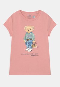 Rosa kortärmad t-shirt med en illustration av en björn i en grön tröja och blå jeans, med vita sneakers och en väska.