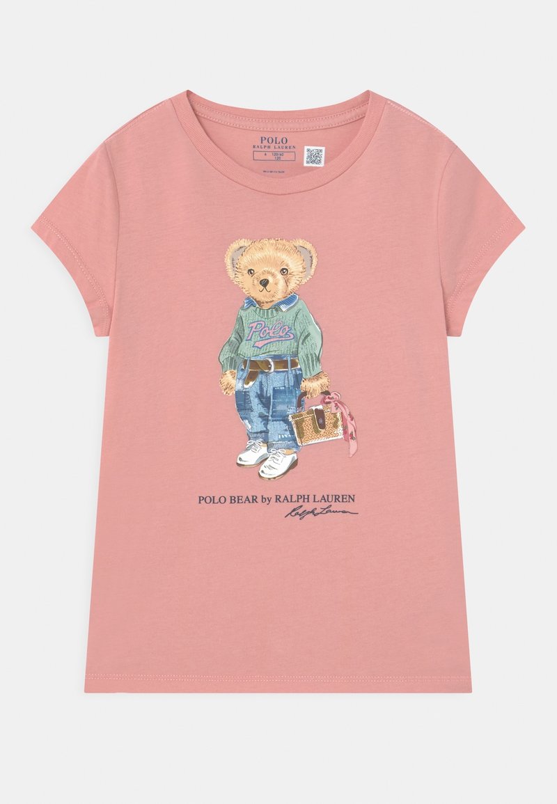 Rosa kortärmad t-shirt med en illustration av en björn i en grön tröja och blå jeans, med vita sneakers och en väska.