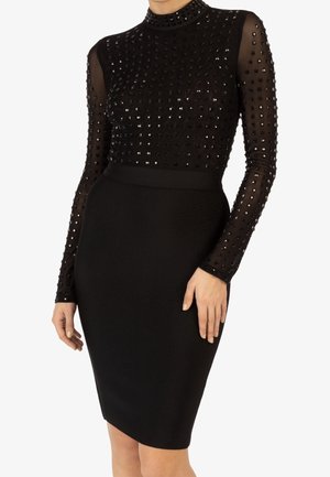 Vestido negro ajustado con mangas largas transparentes y cuerpo superior adornado con tachuelas cuadradas negras, escote alto, falda hasta la rodilla.