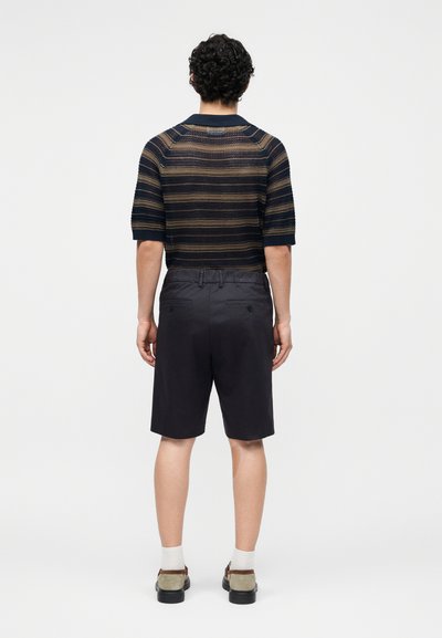Homme vu de dos portant un pull à manches courtes rayé bleu marine et beige, un short noir, des chaussettes blanches et des chaussures beiges sur fond uni.
