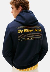 Giovane uomo con capelli ricci che indossa un hoodie blu navy con grande scritta gialla "The Hilfiger Herald" e una stampa aggiuntiva sul retro.