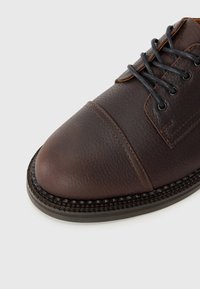 Zapato de cuero marrón con una parte superior texturizada, punta redonda y cordones gris oscuro. Presenta costuras decorativas y un ribete trenzado alrededor de la suela.