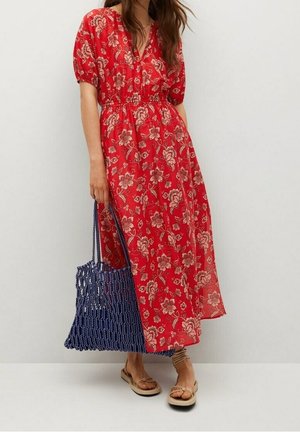 Kobieta w czerwonej sukience midi w kwiaty z bufkami, trzymająca niebieską tkana torbę typu tote i nosząca beżowe płaskie sandały.