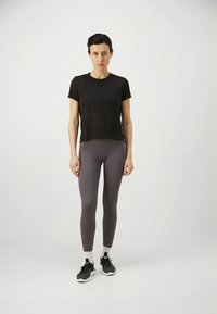 Črna majica s kratkimi rokavi v kombinaciji s sivimi legicami do kolen, tesno prilegajočimi se telesu. Model nosi črne športne copate z belimi poudarki.