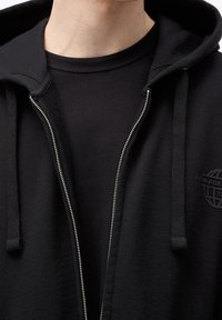 Gros plan sur une personne portant un sweat à capuche noir à fermeture éclair partiellement ouverte, laissant entrevoir un t-shirt noir à col rond en dessous.