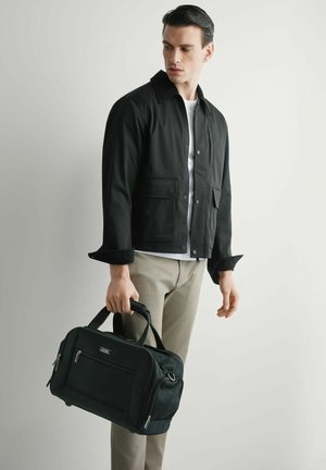 Mężczyzna w czarnej kurtce i beżowych spodniach trzymający czarną torbę podróżną, patrzący w dół w lewo na tle jednolitego jasnego tła.