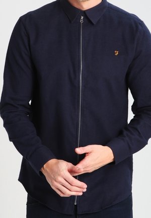 Mężczyzna ubrany w granatową koszulę z długim rękawem, zapinaną na zamek z przodu, z małym żółtym wyszytym na piersi literą "F", z złączonymi dłońmi.