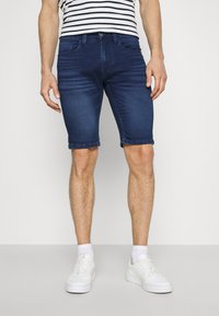 Hombre vistiendo pantalones cortos de mezclilla azul oscuro, zapatillas blancas, calcetines blancos y una camisa de manga corta a rayas blancas y negras, de pie sobre un suelo llano.