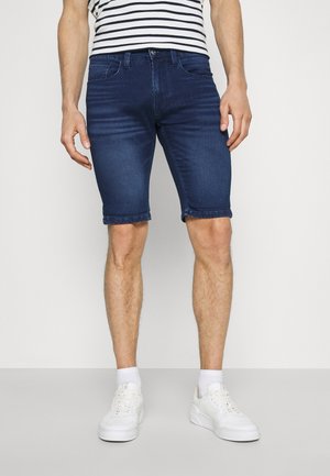 Homme portant un short en denim bleu foncé, des baskets blanches, des chaussettes blanches et une chemise à manches courtes blanc et noir à rayures, se tenant sur un sol uni.