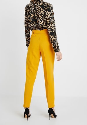 Mujer con pantalones de talle alto color mostaza, blusa de manga larga con estampado de leopardo y zapatos de tacón negro, de espaldas.