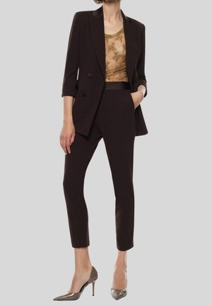 Mujer con un blazer marrón oscuro a medida y pantalones cortos, con una blusa floral beige transparente y zapatos de tacón grises.