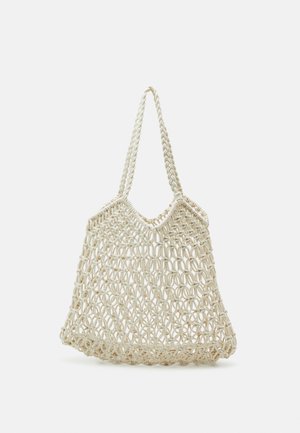 Crèmekleurige macramé draagtas met gevlochten handvatten en open netpatroon, hangend tegen een effen lichte achtergrond.