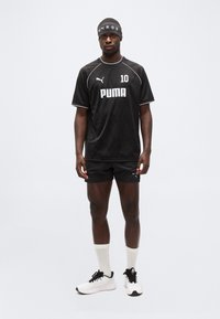 Άνδρας που στέκεται φορώντας μαύρη αθλητική φόρμα Puma με το νούμερο 10, μαύρη κορδέλα στο κεφάλι, λευκές κάλτσες και λευκά αθλητικά παπούτσια, μπροστά σε απλό φόντο.