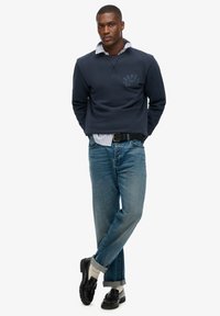 Marineblauwe sweatshirt met logodetail, gedragen over een lichtblauw gestreept overhemd. Gecombineerd met lichtblauwe spijkerbroek en zwarte schoenen met sokken.