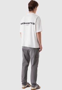 White t-shirt with "PROHIBITED" printed in black on the back, paired with gray cargo pants and beige sneakers with blue stripes.
