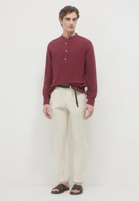Un giovane uomo indossa una camicia rossa con maniche lunghe e bottoni, pantaloni crema con una cintura intrecciata e sandali scuri intrecciati, mentre sta in piedi su uno sfondo bianco.