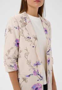 Blazer à imprimé floral en beige clair. Présente des fleurs violettes et bleues avec des contours noirs. Revers crantés, fermeture à un bouton et manches retroussées.
